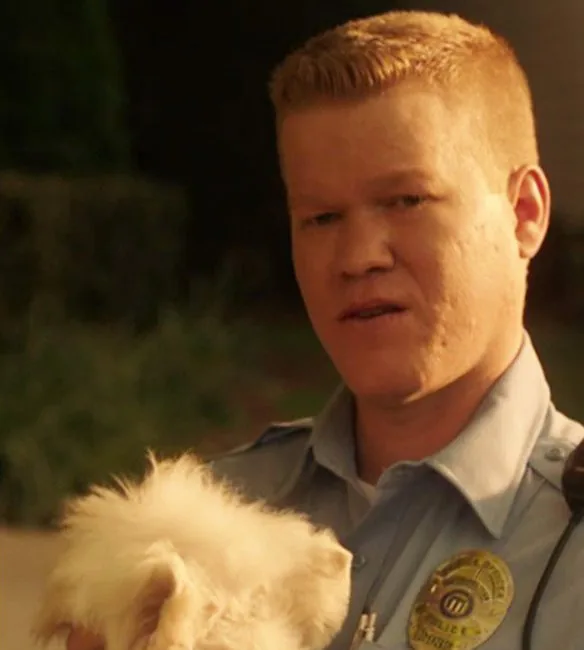 Jesse Plemons as Gary, the deadpan cop neighbor from Game Night (2018), holding his fluffy white dog and asking: How can that be profitable for Frito-Lay?